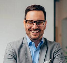 Portrait of young happy businessman wearing grey suit and blue shirt standing in his office and smiling with arms crossed